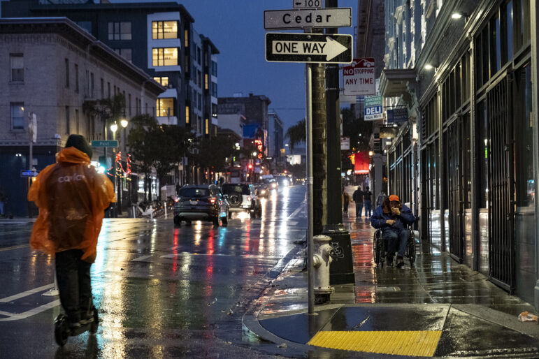 A person wearing an orange rain pancho stands riding a motorized scooter down a rainy street away from the person taking the photo. Cars have their headlights on because it is early evening, and there are lights in the windows of the mid-rise buildings lining the street on both sides. A person in a wheelchair heads down the sidewalk on the right side of the frame toward the person taking the photo.