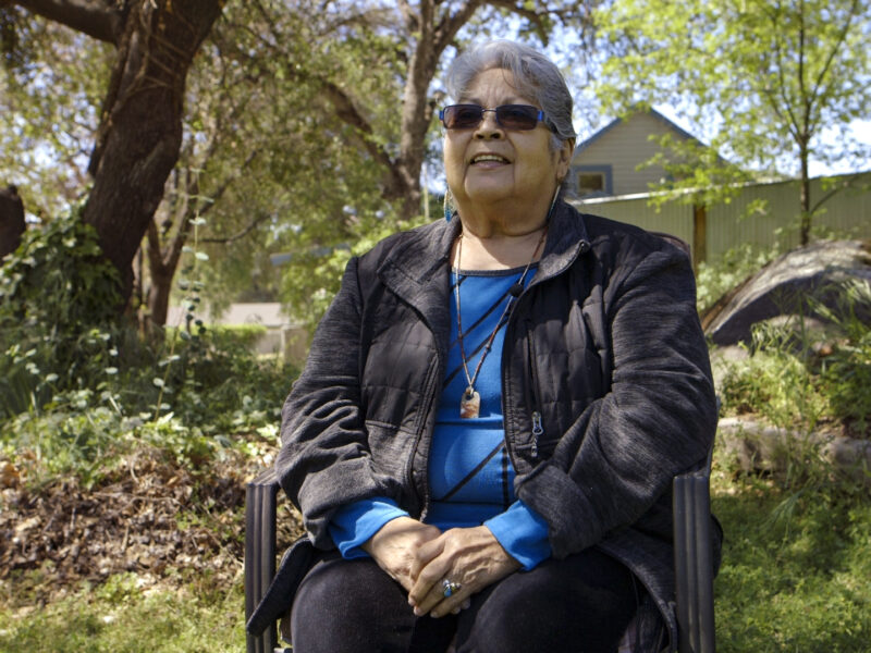 A Native American woman in a blue shirt and black jacket sits on a chair in a forested area near a house.