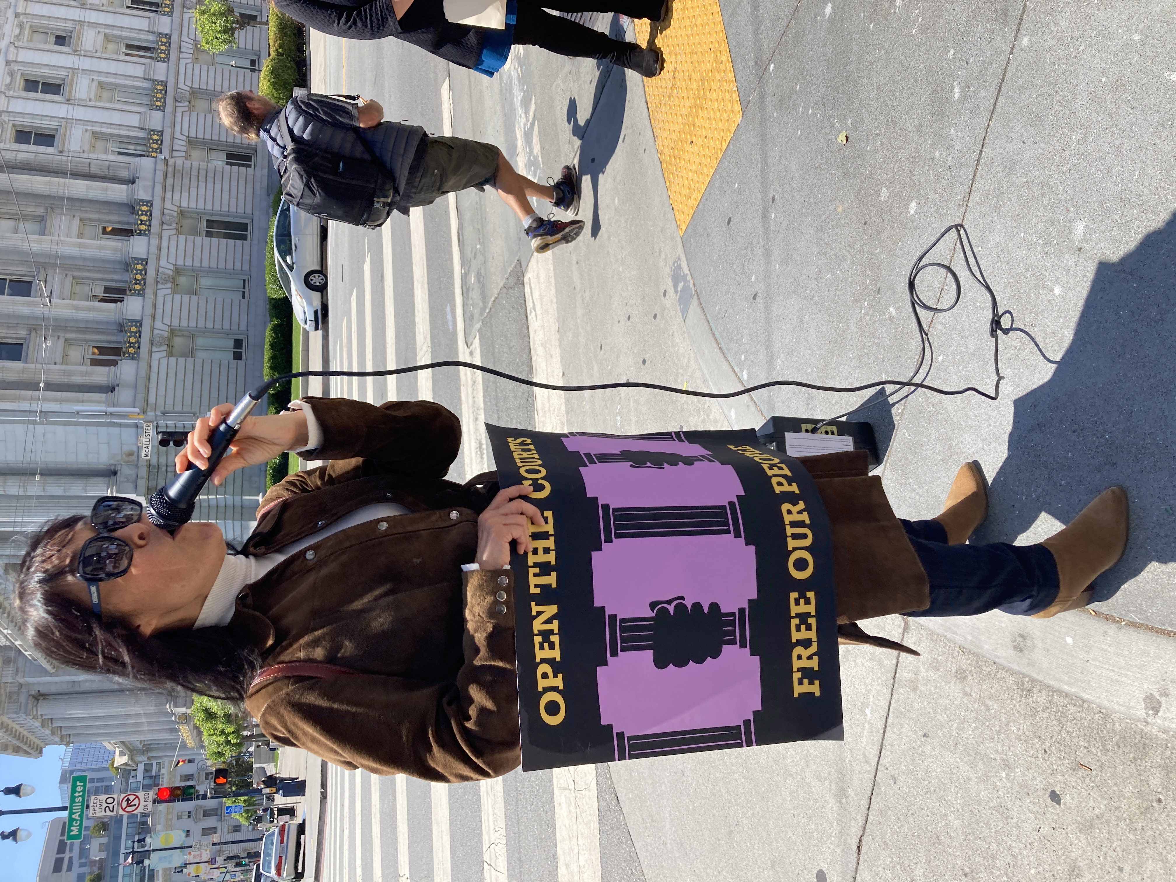 A woman wearing large sunglasses and a long brown coat stands on a street corner and speaks into a microphone while holding a sign that reads "open the courts, free our people."
