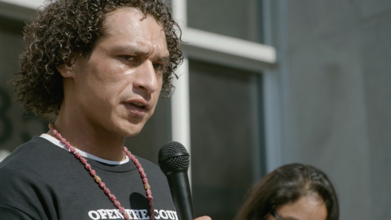 This is a closeup image of a man with brown curly hair wearing a black T-shirt and a necklace with red carved beads speaking into a microphone.