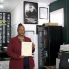 An African American woman in a burgundy jacket and blue pants is standing in an office space with several posters on the wall behind her. She holds a cream-colored booklet with the words "DRAFT San Francisco Reparations Plan" written across the top.
